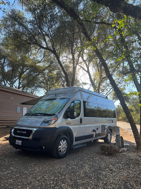 Silver Ram Telaro RV rental parked at a shaded campsite, hooked up. A cabin is nearby, surrounded by lush trees.