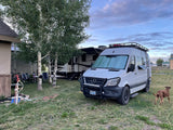 A white Mercedes Sprinter van RV rental and a large travel trailer set up in a grassy campsite with aspen trees and a dog.