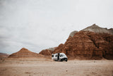 An RV rental in a dramatic desert landscape, parked amidst towering red rock formations under a cloudy sky.
