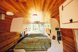 Cozy wood-paneled RV rental interior with a patterned bed, storage, and open back doors revealing a forest.
