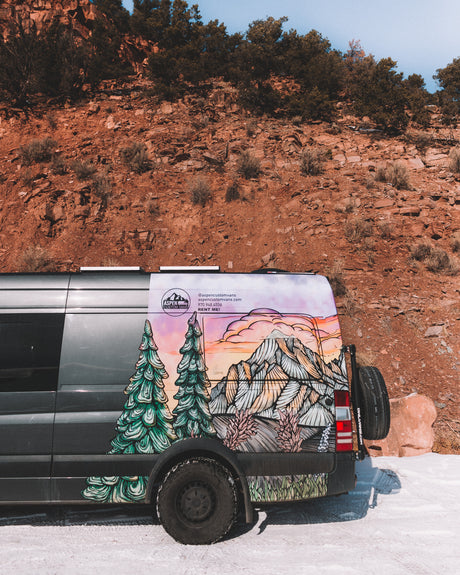 A grey custom rv rental van with a vibrant mountain mural, parked in snow by red rocks. Perfect for scenic adventures.
