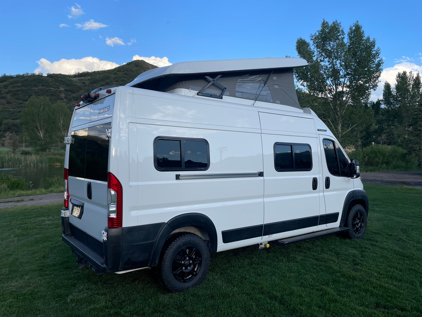 White Winnebago campervan for RV rental, parked on grass by a lake with pop-top open, ideal for nature travel.