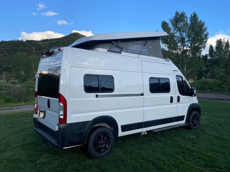 White Winnebago campervan for RV rental, parked on grass by a lake with pop-top open, ideal for nature travel.