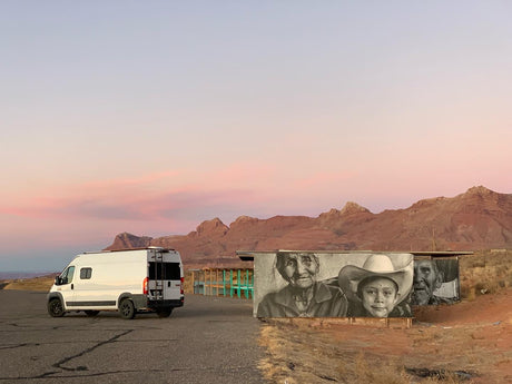 This white RV rental van is parked at sunset, with desert mountains and a building featuring a large mural of faces.