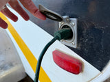 Hand plugs green power cord into RV rental's exterior electrical outlet, with a red taillight below.