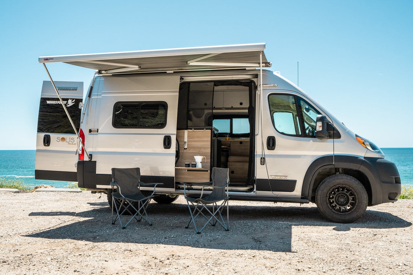 Coastal rv rental: Winnebago Solis Pocket camper van with awning, chairs & coffee setup by the sunny ocean. — photo 7