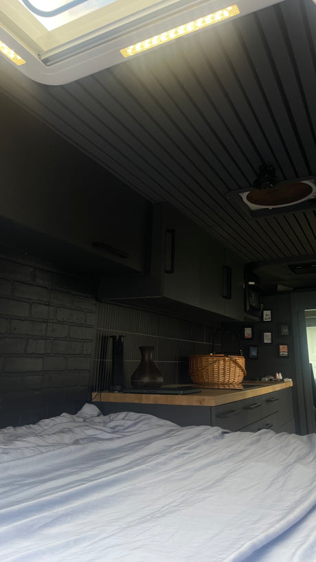 Inside this RV rental: a stylish space with a black brick wall, dark kitchen, comfy bed, and bright skylight. Perfect for mobile life!