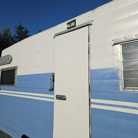 Close-up of a blue and white travel trailer's side, showing its entry door, windows, and a solar light. Consider this RV for rental.