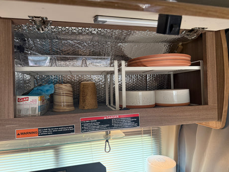 RV rental kitchen cabinet with organized shelves, plates, cups, and safety warnings, insulated with bubble wrap.