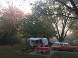 An RV rental and vintage car at a foggy fall campground, featuring a person at a picnic table among colorful trees.