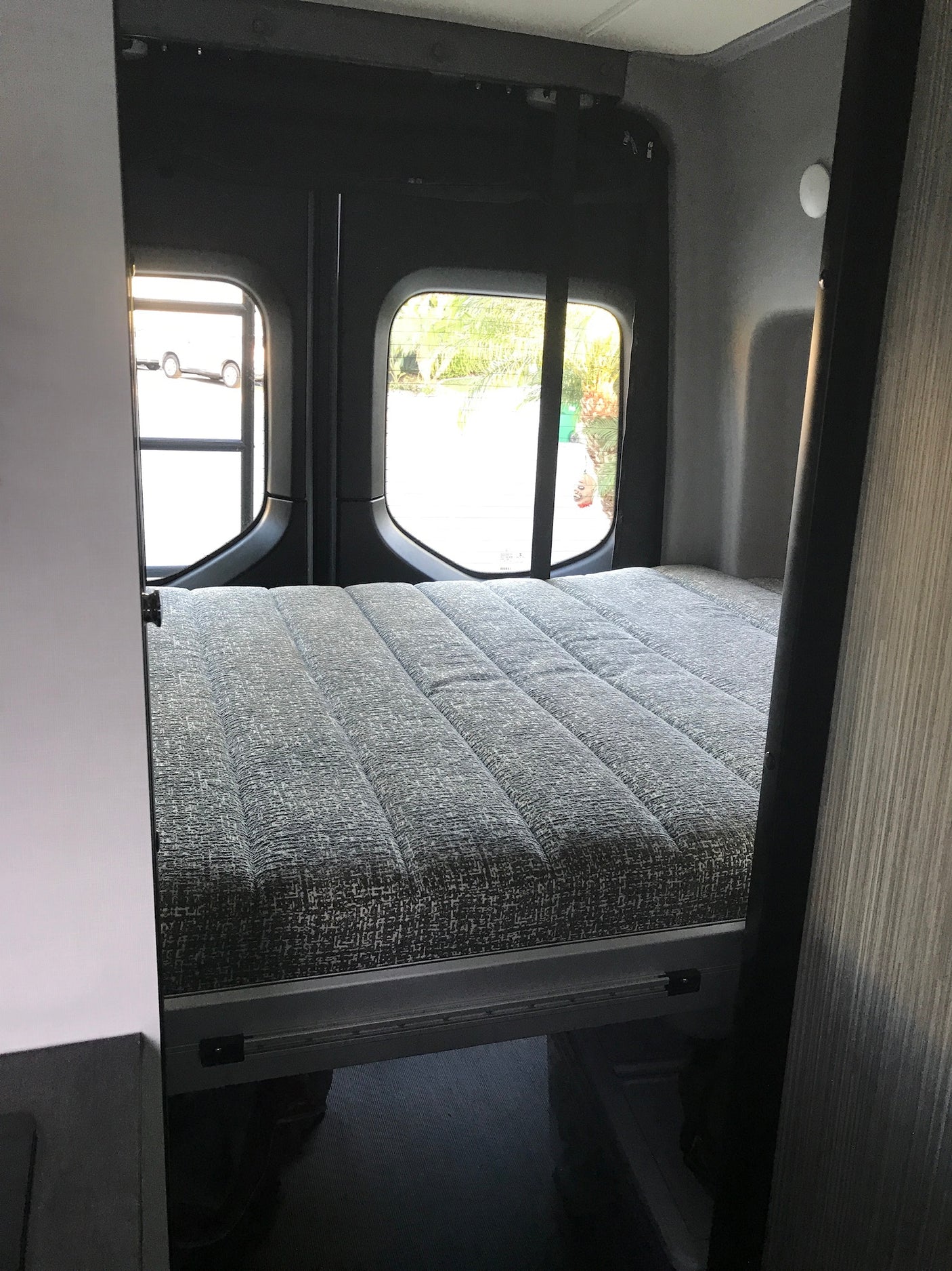 RV rental: Compact sleeping area with a grey textured bed and rear windows, inside a camper van.