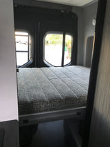RV rental: Compact sleeping area with a grey textured bed and rear windows, inside a camper van.