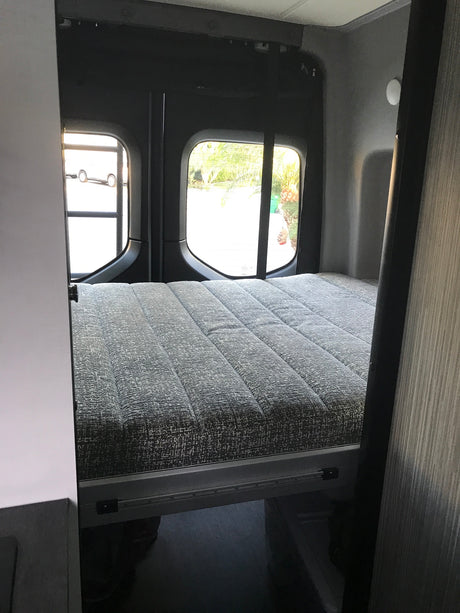 RV rental: Compact sleeping area with a grey textured bed and rear windows, inside a camper van.