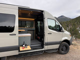 Adventure-ready **RV rental** camper van. Open side door reveals custom interior with kitchen and scenic mountain backdrop.