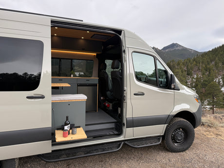 Adventure-ready **RV rental** camper van. Open side door reveals custom interior with kitchen and scenic mountain backdrop.