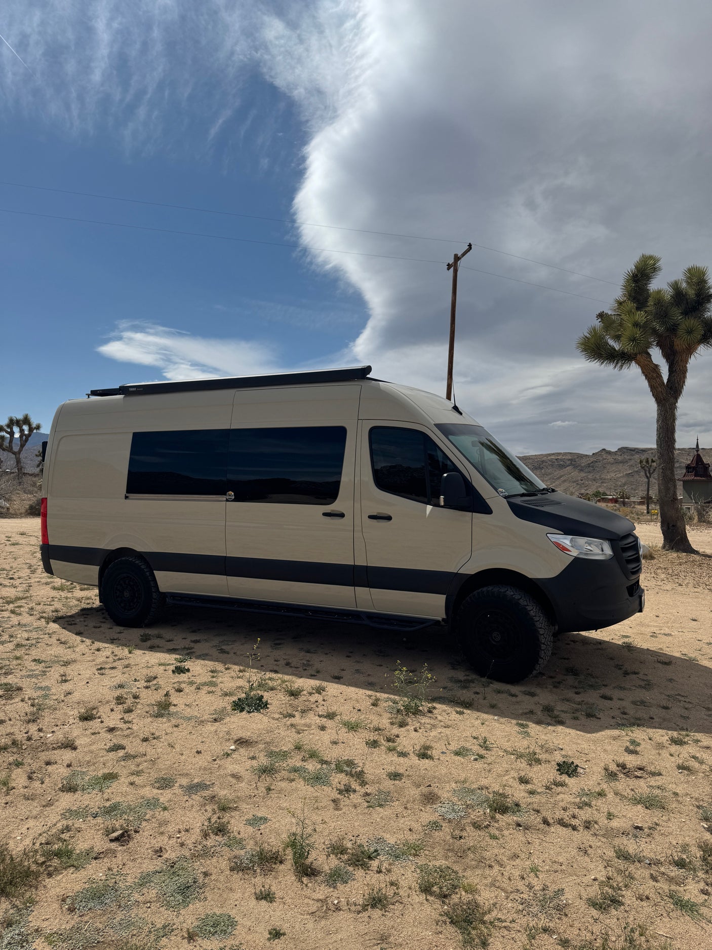 Tan Mercedes Sprinter RV rental van with off-road tires, parked in a desert landscape featuring Joshua trees and clouds.