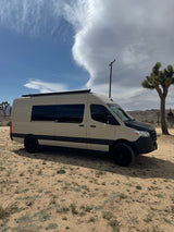 Tan Mercedes Sprinter RV rental van with off-road tires, parked in a desert landscape featuring Joshua trees and clouds.