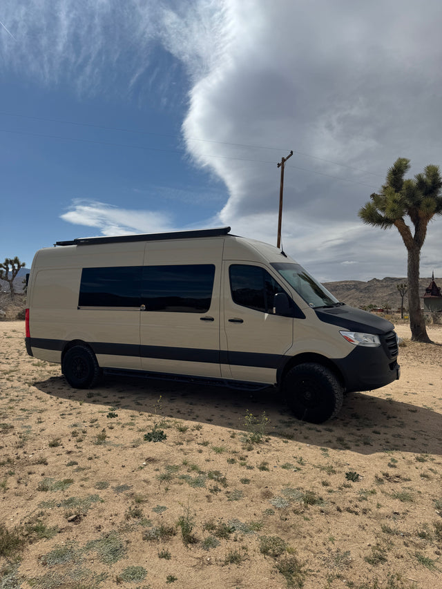Tan Mercedes Sprinter RV rental van with off-road tires, parked in a desert landscape featuring Joshua trees and clouds.