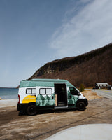 A custom teal & white RV rental van is parked lakeside, with a mountain backdrop. Ideal for scenic adventures.