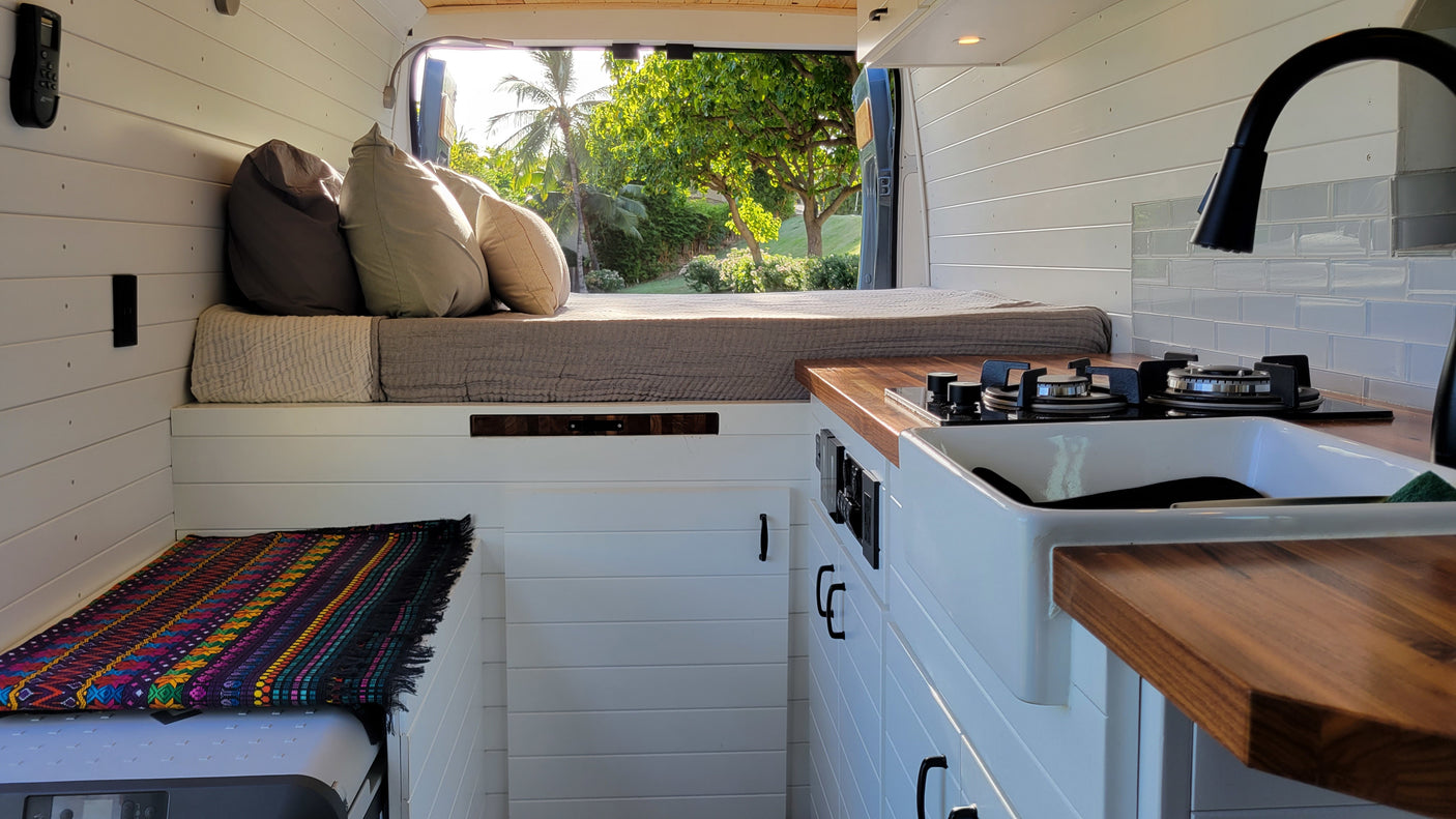 Inside a stylish RV rental van: custom bed, compact kitchen, and lush outdoor views from open doors.