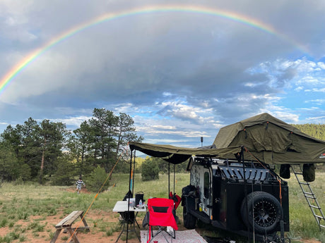 Rainbow overhead at an RV rental campsite: a black overland trailer with rooftop tent, awning, and a picnic setup.