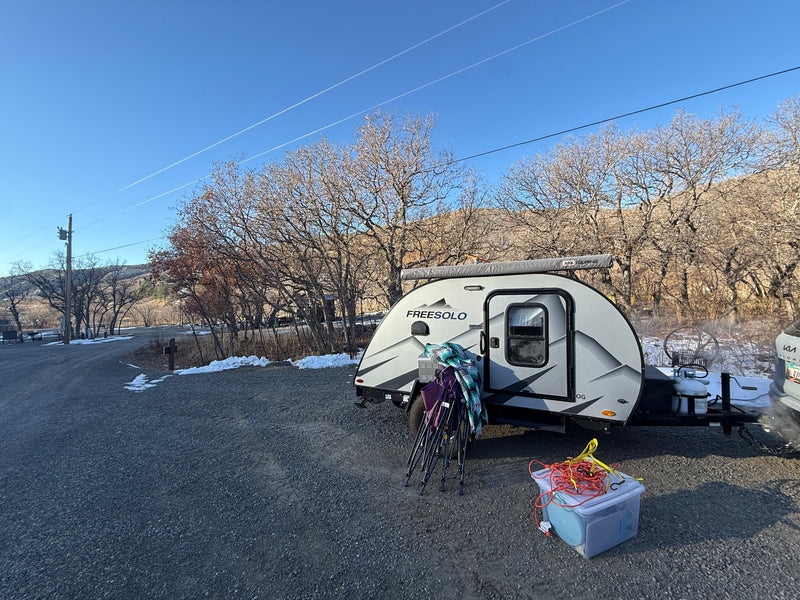 This grey teardrop RV rental is parked at a snowy campsite, surrounded by bare trees and mountains, with camping gear nearby. — photo 6