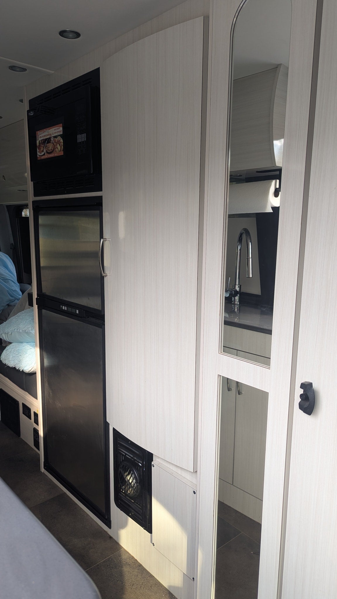This RV rental offers a compact kitchen with microwave, fridge, and light wood cabinets. Mirrored doors reflect the sink area.