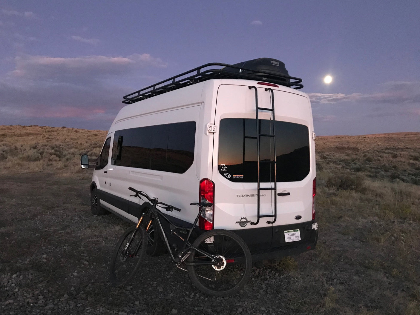 Adventure-ready RV rental: white camper van with bike, roof rack, under a full moon.