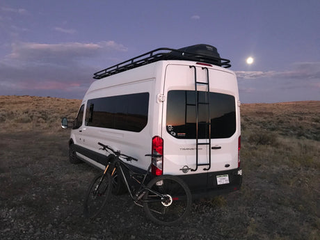 Adventure-ready RV rental: white camper van with bike, roof rack, under a full moon.