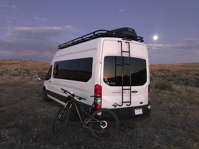 Adventure-ready RV rental: white camper van with bike, roof rack, under a full moon.