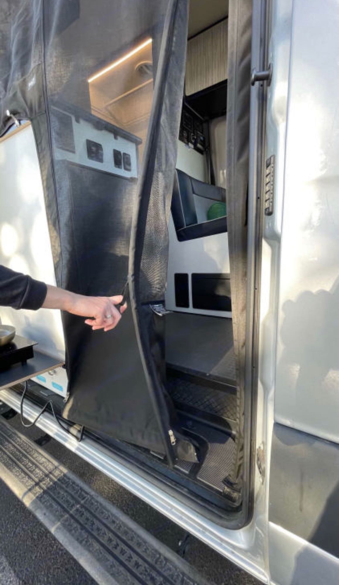 RV rental: Hand opens a black mesh screen door on a camper van, revealing the modern interior with seating & storage.