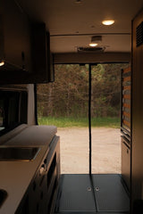 Inside an RV rental, looking out the back screen door to a forest. Features a compact kitchen and living space.