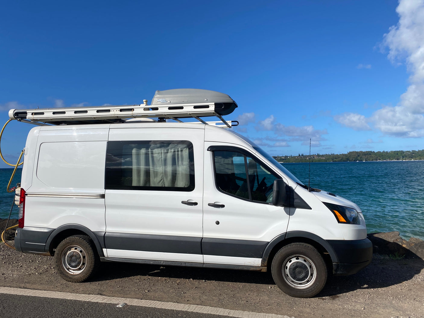 White Ford Transit **RV rental** campervan with roof rack, parked by blue ocean. Ideal for **van life** adventures.