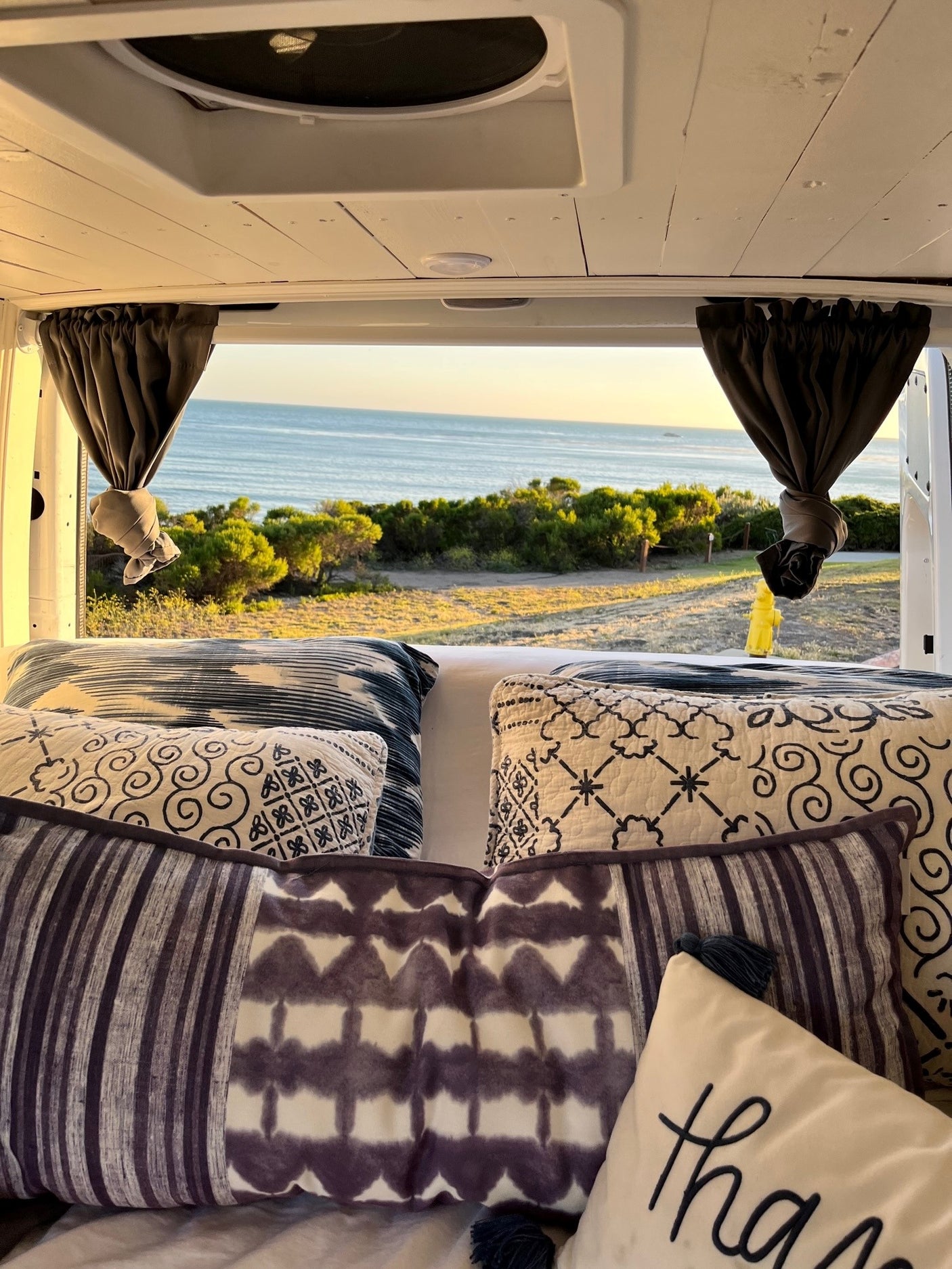 Oceanfront bliss from an RV rental. Cozy bed with patterned pillows offers stunning views of the sea and coastal trees, inviting relaxation.