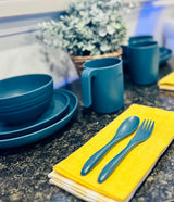 RV rental kitchen supplies: durable blue dishes, mugs, & cutlery on a dark counter with a plant.