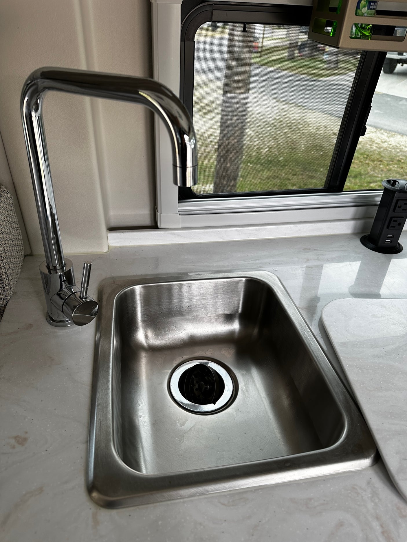 Compact RV rental kitchen features a stainless steel sink, chrome faucet, window with screen, and a convenient pop-up power outlet.