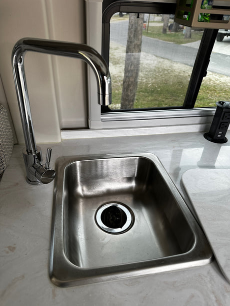 Compact RV rental kitchen features a stainless steel sink, chrome faucet, window with screen, and a convenient pop-up power outlet.