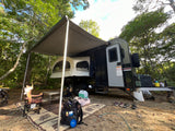 Compact RV rental trailer with awning & pop-out tent bed set up at a shaded forest campsite, with outdoor gear.