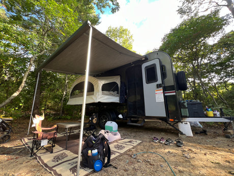 Compact RV rental trailer with awning & pop-out tent bed set up at a shaded forest campsite, with outdoor gear.