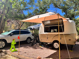 An RV rental camper with its awning deployed, alongside a car at a scenic nature campsite.