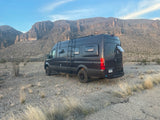 A dark grey Mercedes Sprinter camper van, ideal for RV rental, parked in a vast desert with tall rocky mountains.