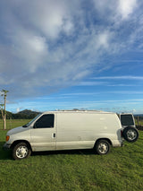 White van parked on green grass, possibly for rv rental. Features include a rear spare tire and muddy sides under a blue, cloudy sky.