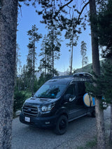 Dark Ford Transit rv rental with paddleboards, nestled among tall pines and mountains. Your perfect nature escape.