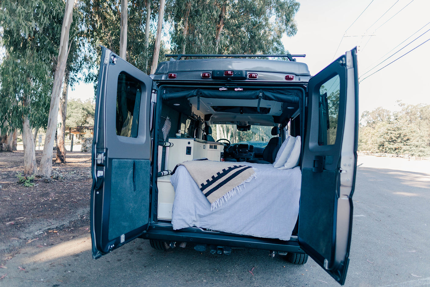 Rear view of an RV rental camper van with open doors, revealing its cozy interior with a bed and kitchenette, parked among trees.