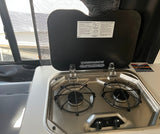 RV rental kitchen showing a compact Suburban two-burner stove with its cover open, displaying instructions.