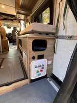 RV rental interior features a functional utility panel for water/power, cozy bed, wood cabinetry, and durable, textured flooring.