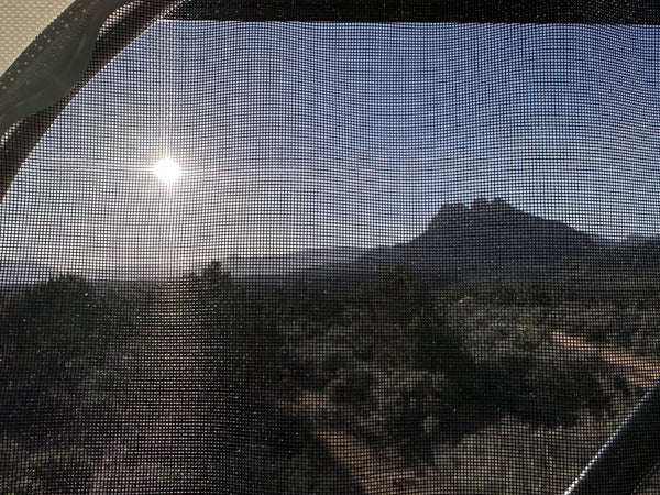 View through an RV rental window screen: bright sun, silhouetted mountain, and wooded landscape. Scenic travel awaits!