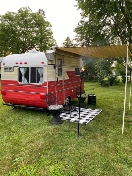 This cozy vintage RV rental features an awning, outdoor chairs, and a lit tiki torch, perfect for a relaxing campsite evening.