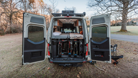 This RV rental's rear doors open to reveal an interior bed and secure bike storage, set in a peaceful outdoor spot.