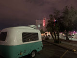 Night view of a retro **rv rental** camper, white & turquoise, parked at Circus Circus Las Vegas. Bright neon sign and trees visible.